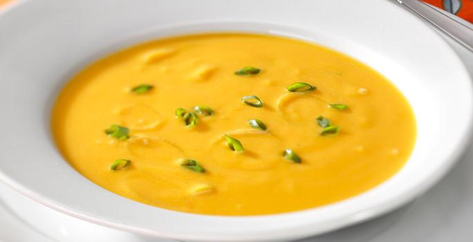 Fotografia em tons de branco e laranja de uma bancada laranja, um supla branco e um prato branco, sobre ele uma sopa de abóbora.