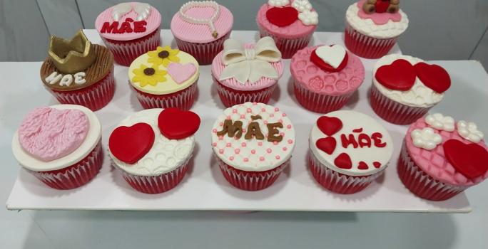 Fotografia em tons de rosa com vários cupcakes ao centro. Cada cupcake possui uma decoração específica, alguns feitos de coração, outros de flores e outros de lacinhos.