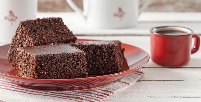 Fotografia em tons de vermelho em uma bancada de madeira clara com um pano listrado em branco e vermelho e um prato vermelho com três pedaços de bolo de cacau com coco. Ao lado, uma xícara vermelha de café e ao fundo, dois bules brancos.