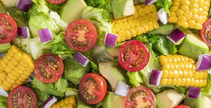 Fotografia vista de cima de uma salada mista com pequenos pedaços da espiga de milho, tomates cereja, cebola roxa, alface e chuchu. A salada está dentro de um recipiente de vidro branco, sobre uma toalha de mesa branca. Ao redor, tomates, abacate e limão.