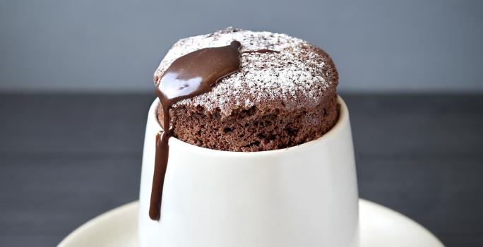 Fotografia de um suflê de café com NESCAFÉ e açúcar de confeiteiro polvilhado por cima. A sobremesa está em um recipiente de vidro branco alto em cima de um prato pequeno branco de sobremesa, e uma colher de café apoiada.