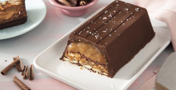 Foto da receita de Barra de chocolate e caramelo crocante. Observa-se uma barra de chocolate cortada em uma travessa grande. Ao lado direito, uma espátula de cortar e, ao esquerdo, um pedaço da torta cortada