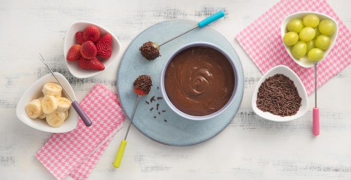 fotografia em tons de rosa e cinza de uma bancada vista de cima, ao centro panos listrados na cor rosa e branco, uma mini tabua azul com um recipiente redondo em cima com chocolate, 4 potes brancos um com granulado e os demais com frutas.