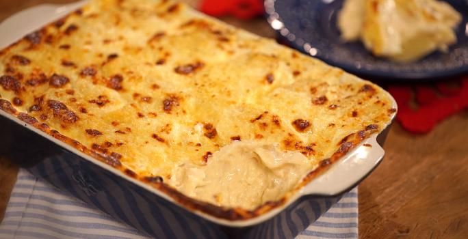 Fotografia em tons de marrom e azul de uma bancada de madeira com um paninho listrado azul e branco, sobre ele um recipiente retangular azul com o gratin. Ao fundo um paninho vermelho e um prato redondo azul com gratin.