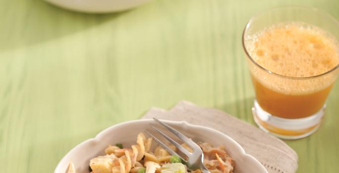 Fotografia em tons de verde em uma bancada de madeira verde com um pano bege, um prato fundo bege com a salada colorida com macarrão, ao lado um copo de vidro com um suco de laranja.