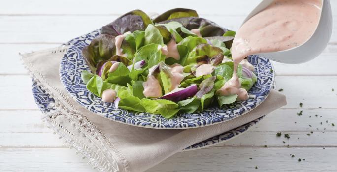 Fotografia em tons de azul, rosa e verde de uma bancada de madeira branca com paninho branco e um prato azul desenhado, sobre ele uma salada de alface com o molho rose e um recipiente despejando o molho sobre a salada.