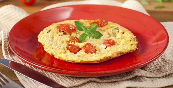 Fotografia em tons de vermelho em uma bancada de madeira, um pano bege, um prato vermelho redondo com a omelete com tomates-cereja em cima dele. Ao fundo, tomatinhos para decorar e ao lado, garfo e faca.
