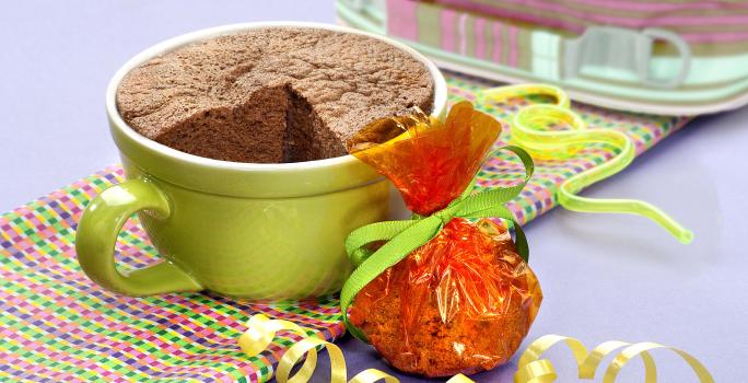 fotografia em tons de cinza, marrom e verde de uma bancada cinza vista de frente, contém um pano em tons de verde, rosa e roxo e uma xicara verde com bolo de chocolate dentro.