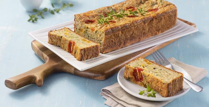 Foto da receita de Tortinha de Legumes. Observa-se uma superfície azul clara, com uma tábua de madeira e, em cima, um prato branco retangular com a torta de legumes inteira, também retangular. Ao lado, um prato branco redondo com um pedaço da torta.