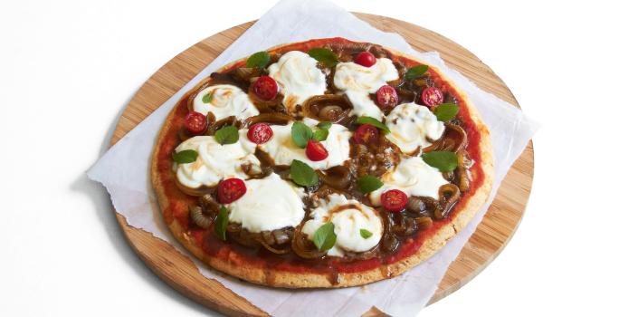 Ao centro, uma tábua de madeira marrom redonda com um pano branco e uma pizza com tomates, cebolas e iogurte por cima.