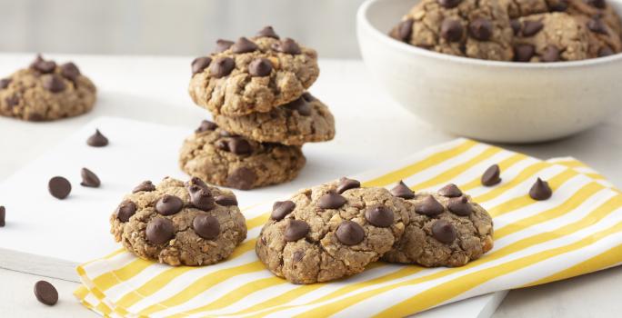 Fotografia em tons de branco e amarelo de uma bancada branca com uma tabua branca e um paninho listrado amarelo e branco sobre ele cookies. Ao fundo um recipiente redondo branco com cookies e gotas de chocolate espalhadas pela mesa.