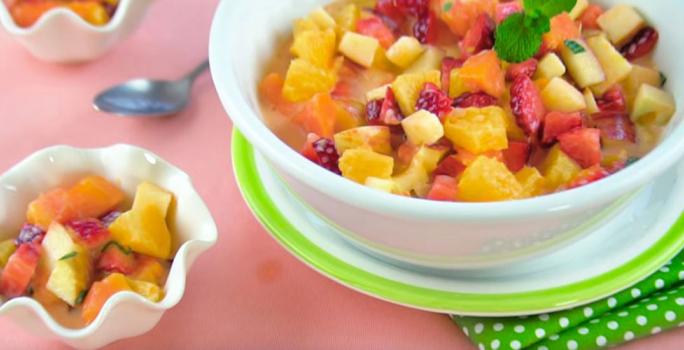 Fotografia em tons de rosa, branco e verde de uma bancada rosa vista de cima, um pano verde com bolinhas brancas e acima um prato branco e verde e por cima do prato um recipiente branco e redondo com frutas. Ao lado um pote branco com frutas.