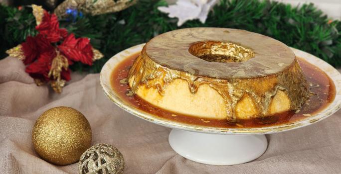 Fotografia em tons de dourado em uma bancada com um pano dourado e enfeites de natal. Ao centro uma bandeja redonda com um pudim de leite Moça e uma calda de caramelo com corante dourado comestível em cima.