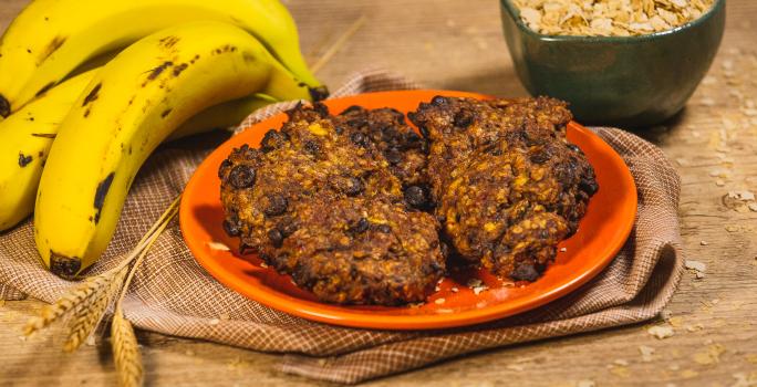 Foto da receita de Cookie Fitness de Banana e Neston, sobre um prato laranja em uma bancada de madeira decorada com mais bananas, trigo e uma caneca com cereais