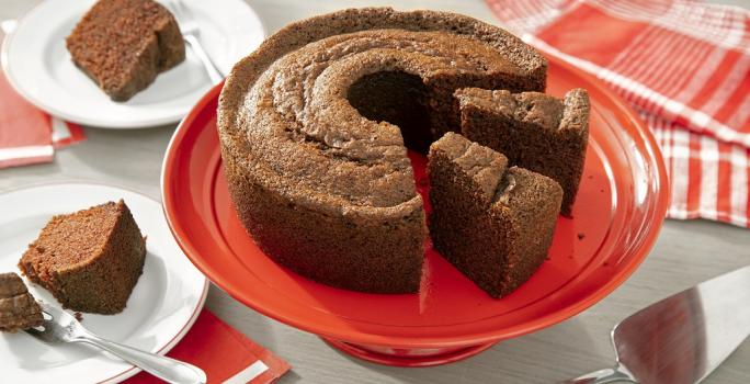 Fotografia em tons de marrom em uma bancada de madeira de cor cinza. Ao centro, uma boleira vermelha contendo o bolo de chocolate. Ao fundo, há um pano vermelho listrado e ao lado, há 2 pires com 2 fatias de bolo.