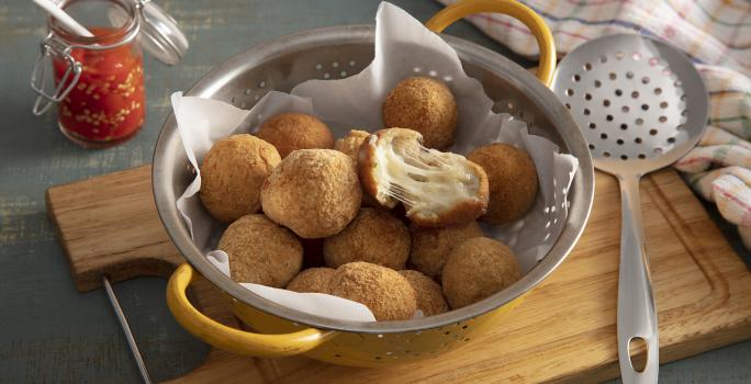 Foto em tons de marrom da receita de bolinho de mandioca (aipim) com queijo servida em diversas porções em um escorredor sobre uma tábua de madeira em cima de uma mesa cinza chumbo. Ao lado há uma escumadeira prateada e um paninho colorido