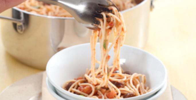 Fotografia em tons de branco e bege, com bowl branco contendo macarrão com molho Putanesca e pegador de massa seguro por uma mão, sobre guardanapo bege e sous plat de inox, panela de inox com macarrão ao lado, tudo sobre bancada bege.