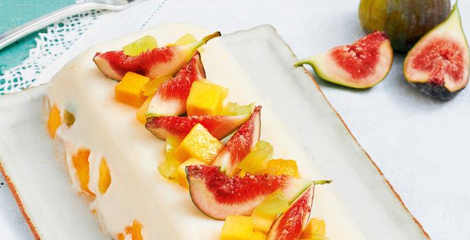 Fotografia em tons de branco e azul claro de uma bancada vista de cima, contém um pano branco, uma travessa branca retangular que contém a sobremesa de gelatina com diversas frutas por cima para decorar.