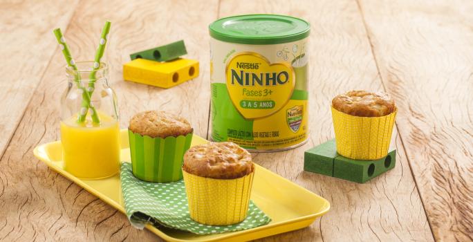 Fotografia em tons de amarelo e verde em uma bancada de madeira clara, um recipiente amarelo com dois cupcakes de maçã e aveia em forminhas amarela e verde. Ao lado, brinquedinhos infantis, uma lata de Ninho Fases e mais um cupacke.