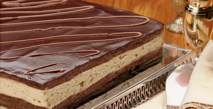 fotografia em tons de marrom de uma bancada marrom vista de frente, em um suporte de alumínio contém a torta com camadas de mousse e chocolate por cima.