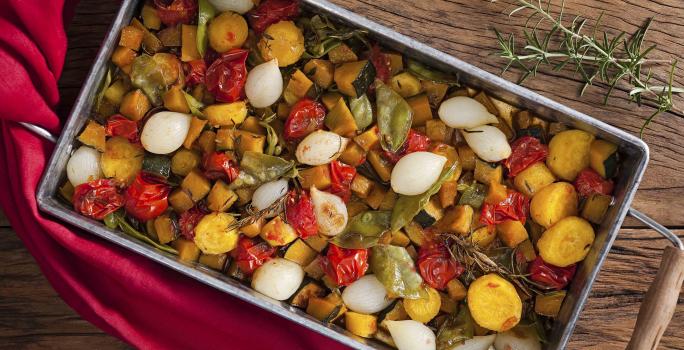 Fotografia de um recipiente de ferro com alças de madeira, dentro estão todos os legumes ficados em cubos assados, e ao lado está um guardanapo de tecido vermelho.