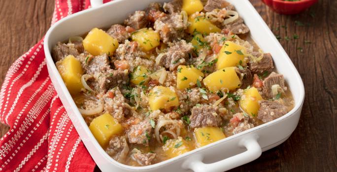 Fotografia em tons de marrom e branco uma bancada vista de cima, ao centro um recipiente quadrado com alças cor branco, dentro dele cubos de carne e pedaços de Mandioquinha e Quinoa. Ao lado um pano listrado na cor vermelho e branco.