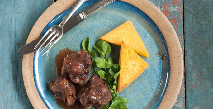 Fotografia em tons de azul em uma bancada de madeira azul com um prato redondo raso rosa claro com azul e a rabada, a polenta e salada de agrião em cima dele.