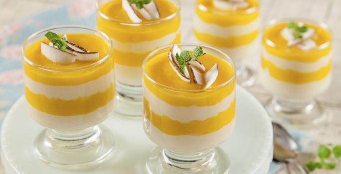 Fotografia em tons de branco e amarelo, com cinco taças contendo camadas de cremes amarelo e branco alternadas, três delas sobre suporte branco, duas colheres ao lado, tudo sobre bancada de cor clara.
