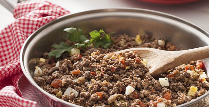 Fotografia em tons de vermelho em uma bancada de madeira com um pano quadriculado em vermelho com uma frigideira com carne moída suculenta.