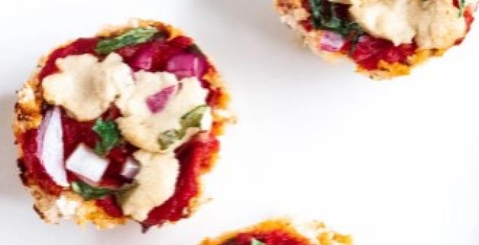 Fotografia em tons de vermelho em uma mesa branca, um prato redondo branco e vários pedaços pequenos de pizza feitas com massa de couve-flor e recheada de molho de tomate, rúcula e queijo mozarela.
