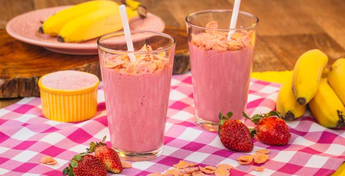 Imagem de uma bancada de madeira com um pano quadriculado em tom de rosa claro, morangos, e cereais espalhados, além de dois copos com uma bebida cor de rosa e canudos.