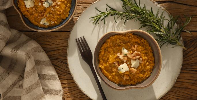 Foto em tons de laranja da receita de risoto de abóbora com gorgonzola servida em um bowl de cerâmica cinza sobre uma base de porcelana branca com um garfo ao lado além de um ramo de alecrim