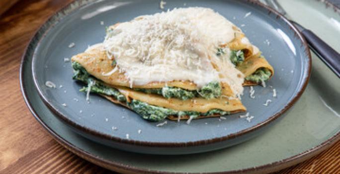 Fotografia de dois crepes de queijo e espinafre dobrados com molho branco por cima e queijo ralado. Os crepes estão em um prato azul raso, que está sore outro prato, um pouco maior, e ao lado, um garfo apoiado.