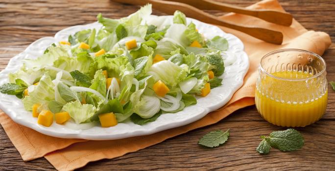 Fotografia em tons de marrom em uma bancada de madeira com um pano laranja e um prato branco oval com as hortaliças. Ao lado, um pegador de salada de madeira e um copo de vidro com suco de laranja.