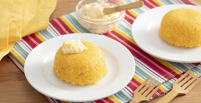 Foto em tons de amarelo da receita de cuscuz de micro-ondas com queijo servida em duas porções sobre pratos brancos de porcelana em cima de uma toalhinha colorida com garfos de madeira e um vidro com queijo ao lado