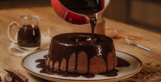 Fotografia em tons de vermelho em uma mesa de madeira com um prato branco com o bolo de chocolate em cima e uma calda de chocolate por cima. Uma panela vermelha em cima do bolo derramando a calda de chocolate.