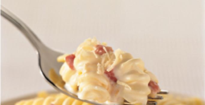 Fotografia em tons de amarelo de um garfo prateado com uma porção de macarrão parafuso cremoso com bacon sobre um pote com macarrão. Fotografia em tons de amarelo de um garfo prateado com uma porção de macarrão parafuso cremoso com bacon sobre um pote com macarrão.