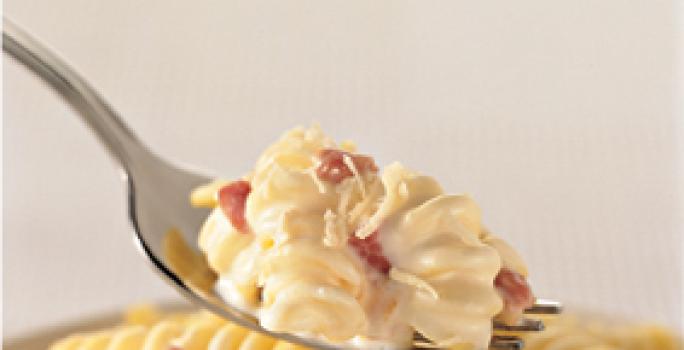 Fotografia em tons de amarelo de um garfo prateado com uma porção de macarrão parafuso cremoso com bacon sobre um pote com macarrão.