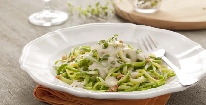 Fotografia em tons de verde e branco, ao centro um prato branco fundo com o espaguete de abobrinha verde dentro com molho branco. Abaixo do prato está um guardanapo de tecido laranja em uma mesa de madeira.