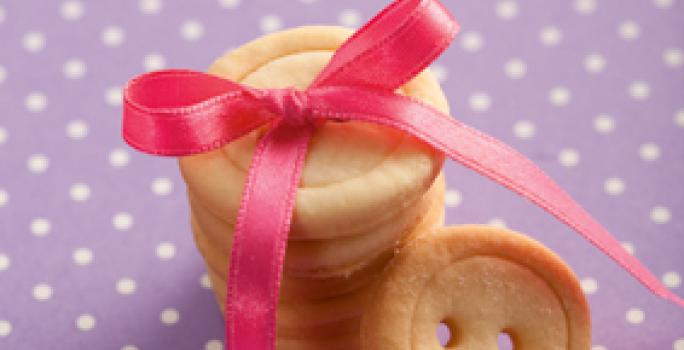 fotografia em tons de roxo, bege e rosa de uma bancada roxa com bolinhas brancas, contém biscoitinhos em formatos de botão com um laço rosa por cima.