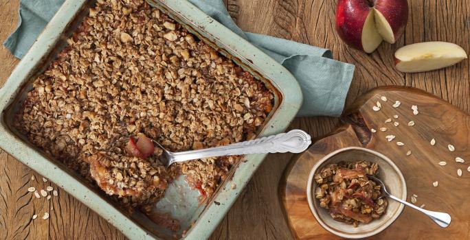 Fotografia em tons de azul em uma mesa de madeira escura com uma travessa retangular azul e o crumble de maçã e aveia dentro. Ao lado, uma tábua de madeira e um potinho com um pedaço do crumble. Ao fundo, uma maçã cortada ao meio.