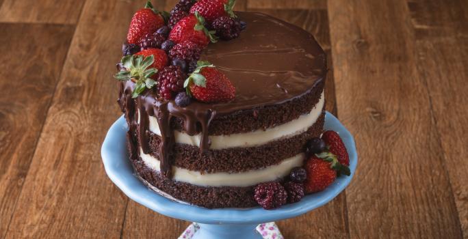 Imagem da receita de Naked Cake de Cacau, decorado em camadas marrons e brancas, com chocolate escorrendo, decorado com frutas vermelhas. O bolo está sobre um prato azul em uma bancada de madeira.