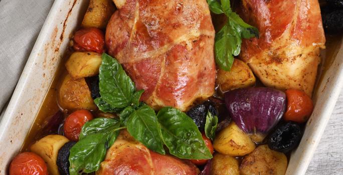 Fotografia vista de cima de três peitos de frango assados e recheados com maçã e ameixa no molho de laranja, e ao redor, vegetais assados que estão dentro de uma travessa de vidro na cor branca. O recipiente está sobre um pano cinza.