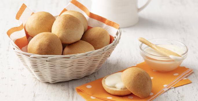 Fotografia em tons de laranja e branco de uma bancada branca com um paninho laranja, sobre ele um pãozinho aberto recheado e um recipiente de vidro com requeijão. Ao lado uma cesta com um paninho laranja e branco com os pães.