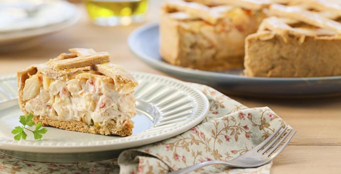 Fotografia em tons de bege em uma mesa de madeira com um pano florido em bege e vermelho e um prato branco apoiado nele com um pedaço de torta de palmito e alho-poró. Ao fundo, um prato grande com a torta cortada ao meio.