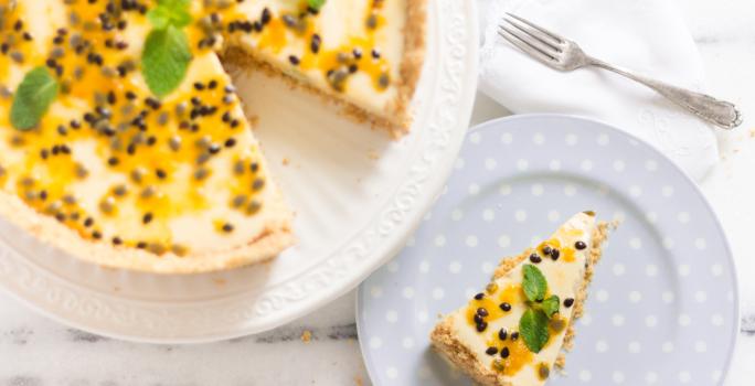 fotografia em tons de branco e amarelo de uma bancada branca vista de cima. Contém um prato branco redondo com uma torta com mousse de maracujá e sementes para decorar, e ao lado um prato redondo azul com a fatia da torta.