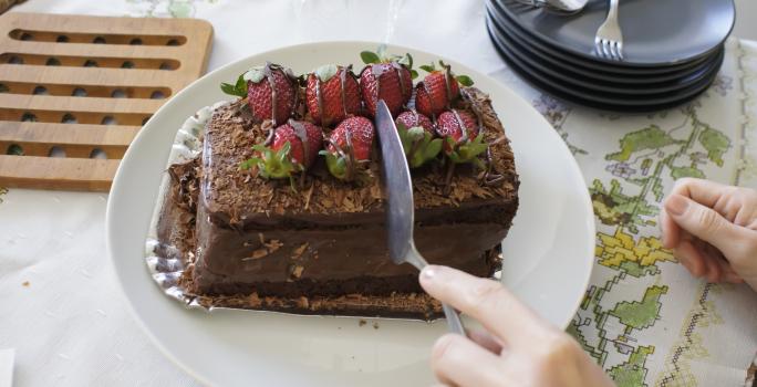 Fotografia de uma mão cortando um bolo de chocolate recheado com morango que está em um prato branco. Ao fundo, alguns pratos pretos empilhados, e ao lado, uma decoração em madeira para apoiar bolo ou pães. Tudo está sobre uma toalha de mesa branca.