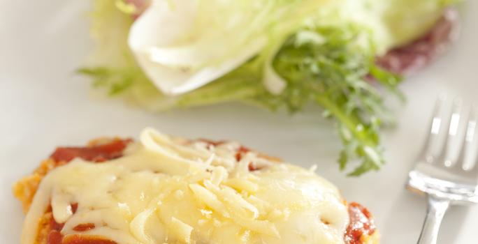 Fotografia em tons de marrom em uma bancada de madeira com uma toalha cinza, um sousplat marrom, um prato branco redondo com o filé à parmegiana acompanhado de salada. Ao fundo, um potinho de azeite.