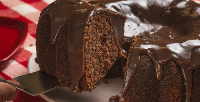 Foto bem aproximada de um bolo de chocolate com uma fatia sendo retirada. O bolo está num prato branco e coberto com Brigadeiro e o prato está sobre uma toalha xadrez vermelho e branco.