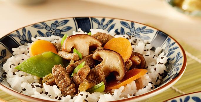 fotografia em tons de branco de bege de uma bancada bege vista de frente. Contém dois recipientes redondos em tons de branco e azul com arroz, cenouras fatiadas e shitake, ao lado o hashi para servir e ao fundo uma xicara branca.